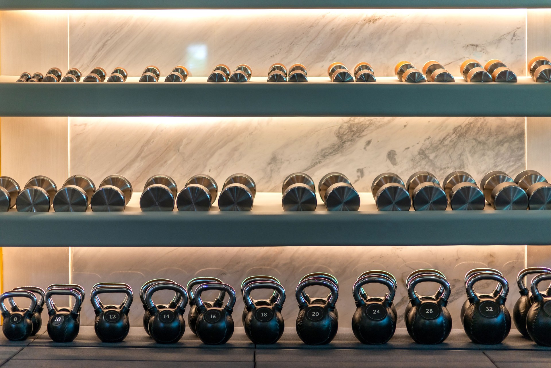 Wide range of choices. Kettlebells weights and dumbbells lying in a row on shelves in stylish workout gym. Marble background. Sport, healthy lifestyle concept. Horizontal shot.